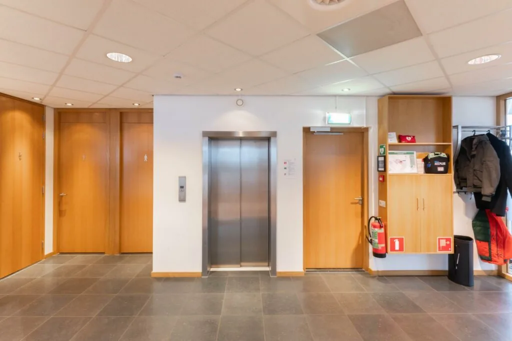 Hal met lift, drie houten deuren, brandblusser en kapstok in een kantoorgebouw aan de 1e Hogeweg.